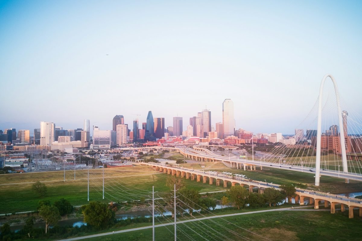 Downtown Dallas skyline at sunset seen across the Trinity River, with the Margaret Hunt Hill Bridge arch
